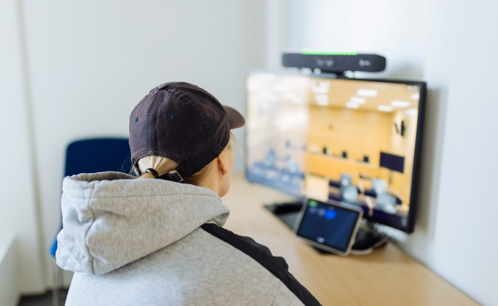 Henkilö istuu pöydän ääressä. Pöydällä on näyttö, jossa näkyy videokuvaa oikeussalista.