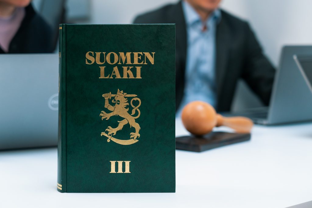 Pöydällä on lakikirja ja nuija. Pöydän takana istuu kaksi henkilöä tietokoneiden ääressä.
