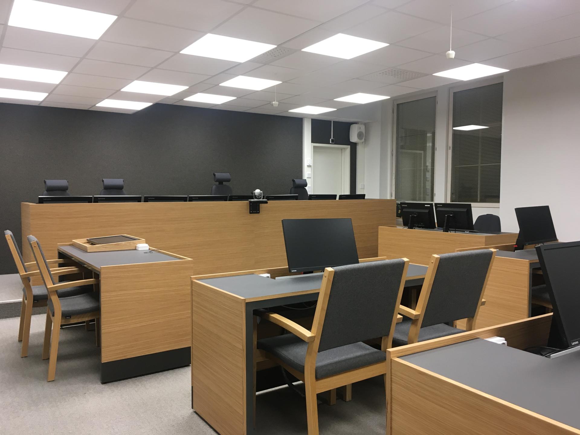 Kuvassa näkyy oikeuden jäsenten pitkä pöytä, syyttäjän ja todistajan pöydät sekä asianosaisten pöydät ja pöydillä olevat näyttöruudut sähköisen materiaalin esitystä varten.